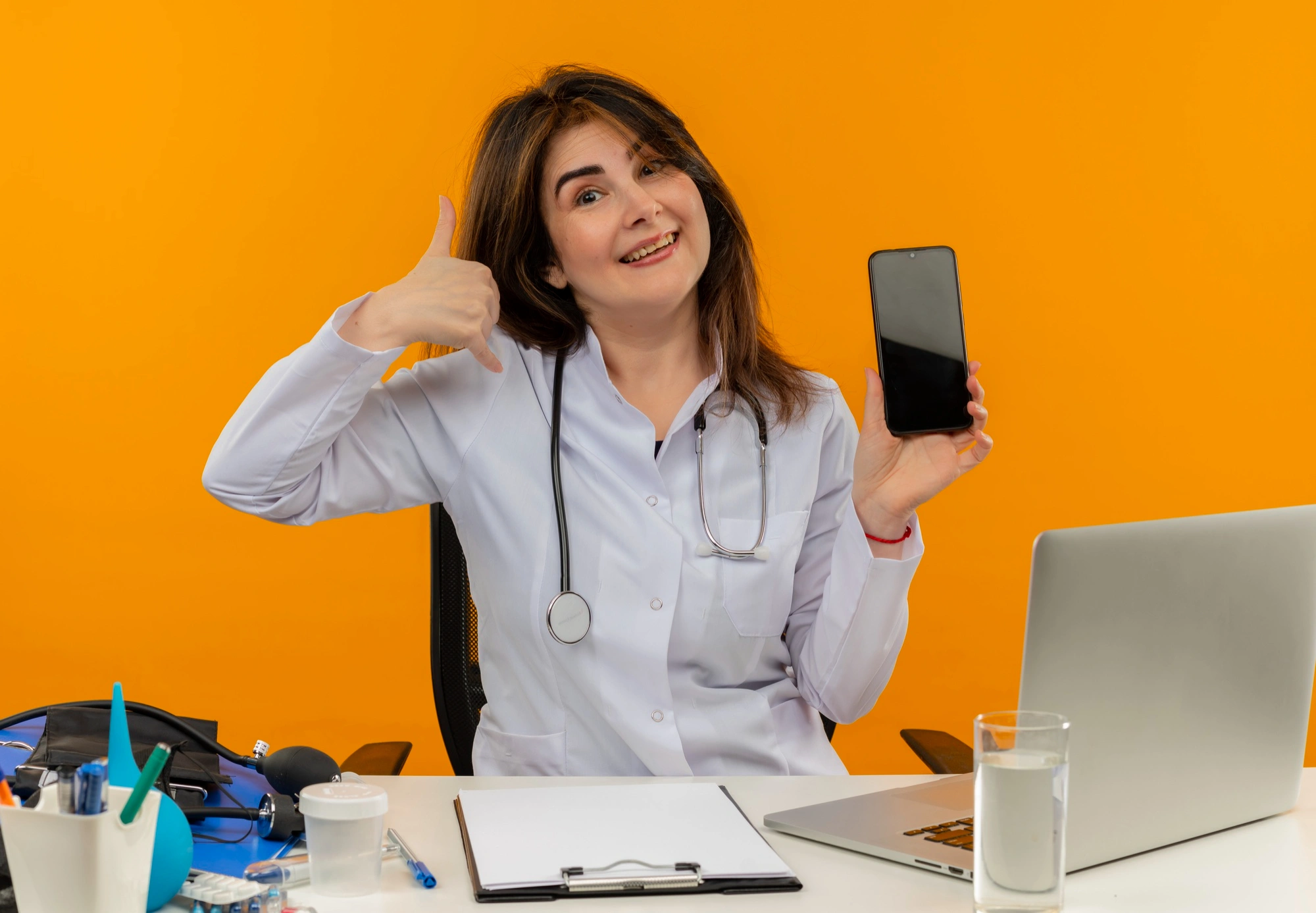 Female doctor in a white coat and stethoscope sitting at a desk, smiling and showing a blank smartphone screen, suggesting telemedicine or online booking.