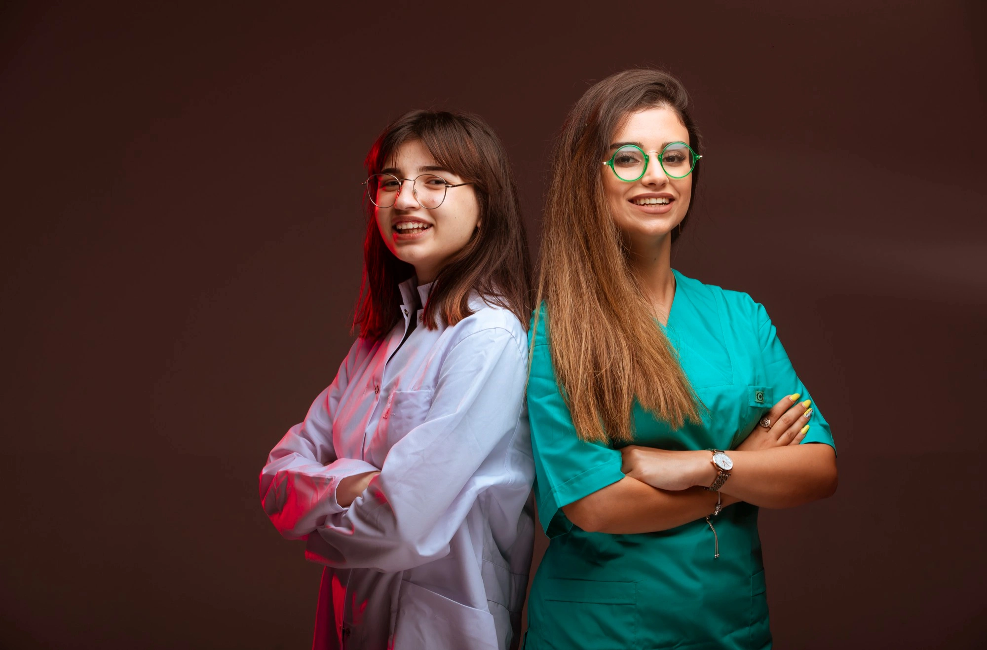 Two smiling female medical or dental professionals standing back-to-back, one in a white coat and one in green scrubs, symbolizing teamwork and expertise.